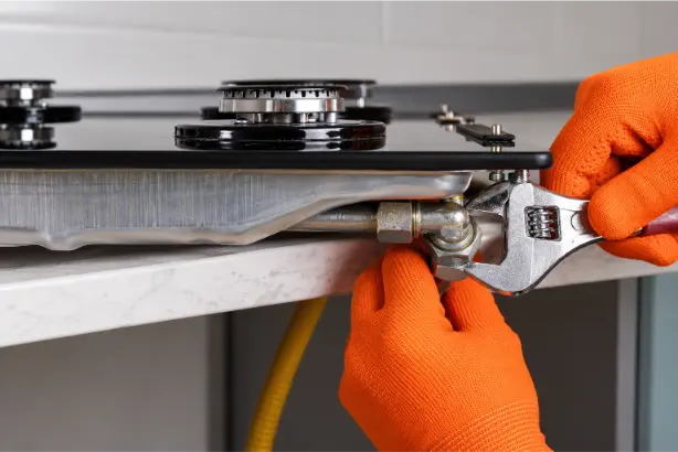 Gas engineer wearing orange protective gloves tightening a gas hob connection with an adjustable wrench beneath a black glass cooktop.
