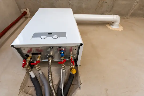 White wall-mounted boiler with exposed pipe connections, red isolation valves and flue in an unfinished utility room.