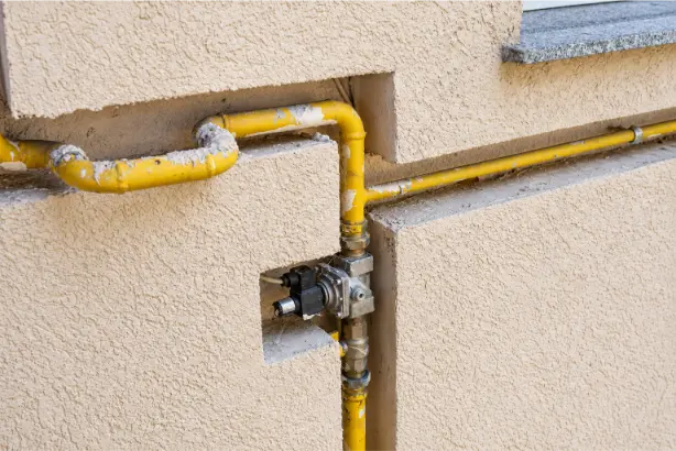 Yellow external gas pipework with a metal gas regulator and fittings mounted in a recessed section of a beige rendered wall.